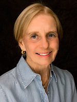 Photographic portrait of Jane Healy wearing a denim jean shirt. She has blond hair parted near the center and pulled back close to her head and an engaging smile on a black background.