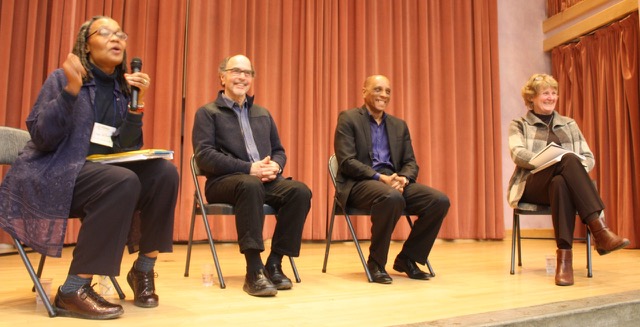 Four people in chairs in a semi-circle on a stage looking out on an unseen audience. One woman is lively and holds a microphone, smiling while speaking. The others are laughing. Panel of Speakers